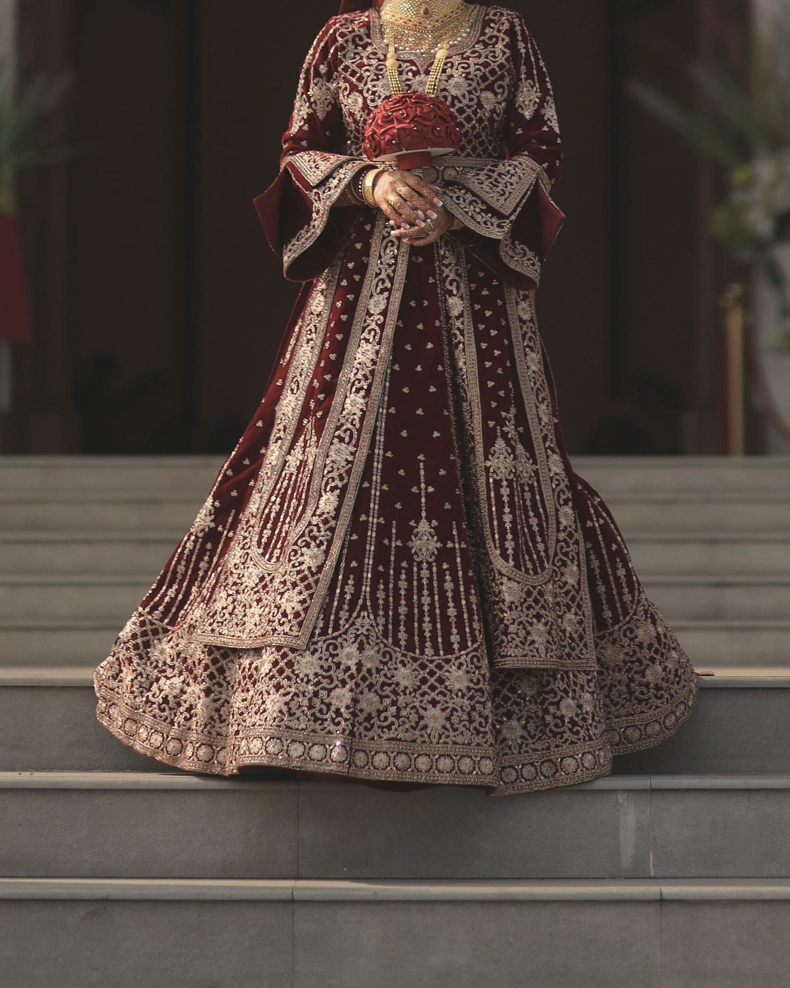 A deep red velvet bridal lehenga with heavy gold zardozi, a bouquet held in hennaed hands, on white steps