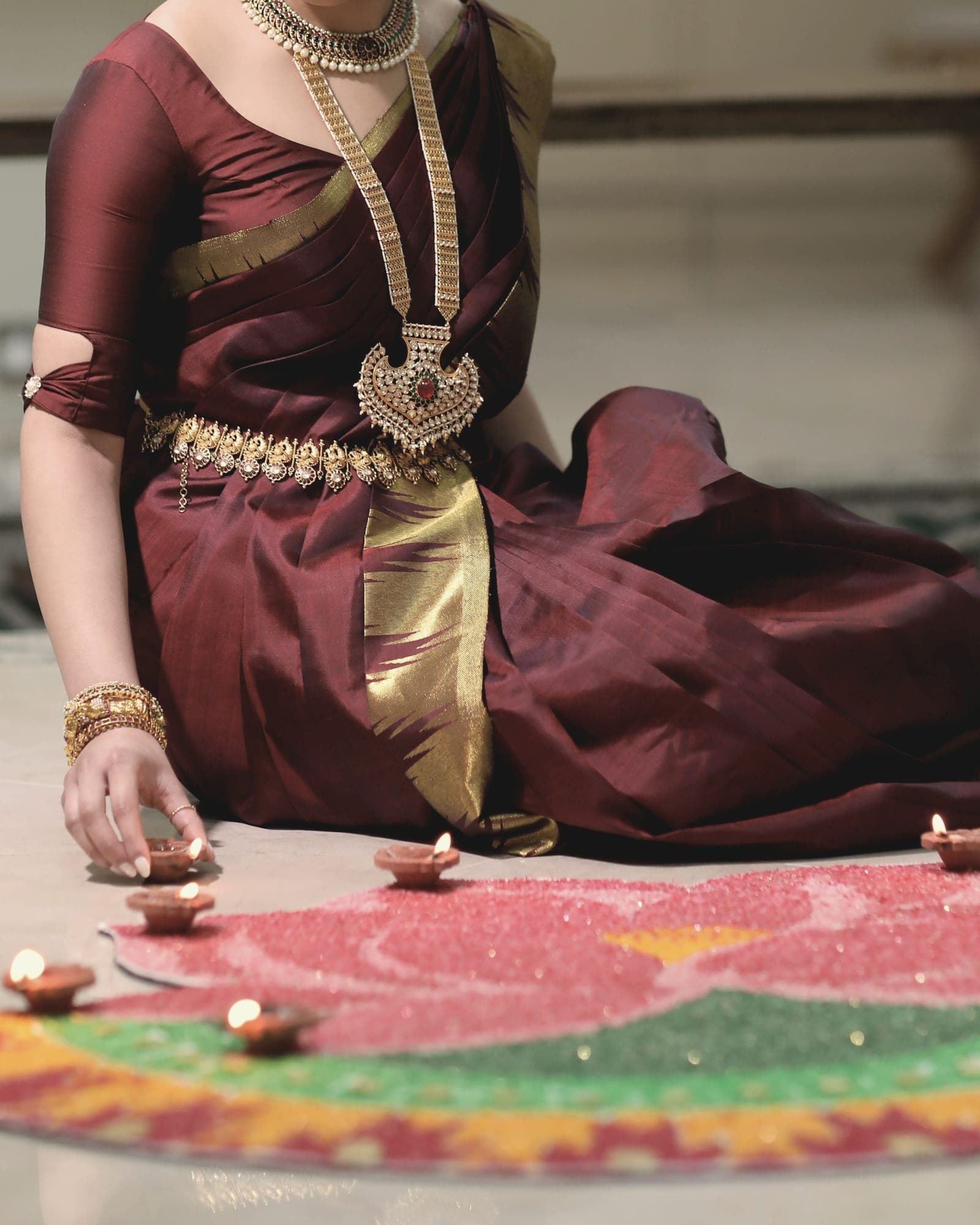 A maroon and gold silk saree with traditional gold jewellery, a hand placing a lit diya beside a rangoli