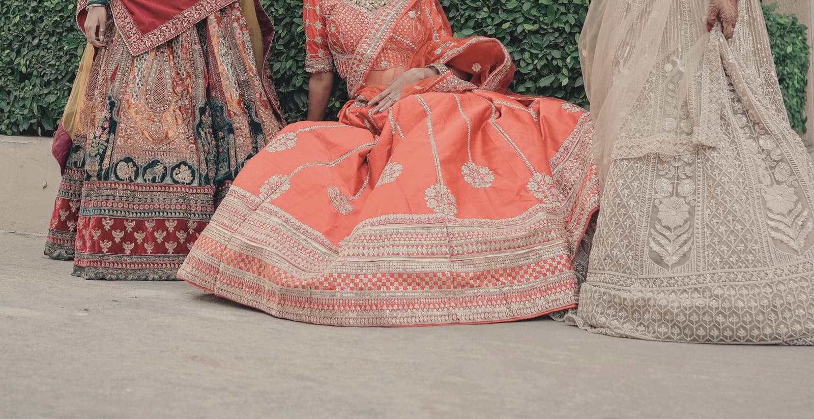 Three bridal lehengas side by side: deep red and gold, coral with silver embroidery, and ivory